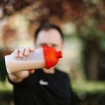 Focus sur un homme tenant un shaker en plastique pour protéines en extérieur avec un arrière-plan flou.