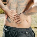 A man with a large tattoo on his back holds his lower back outdoors, showing discomfort. Un homme avec un grand tatouage sur le dos se tient le bas du dos à l'extérieur, montrant de l'inconfort.