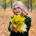 Femme blonde portant des lunettes tenant des feuilles d'automne dans un cadre forestier.