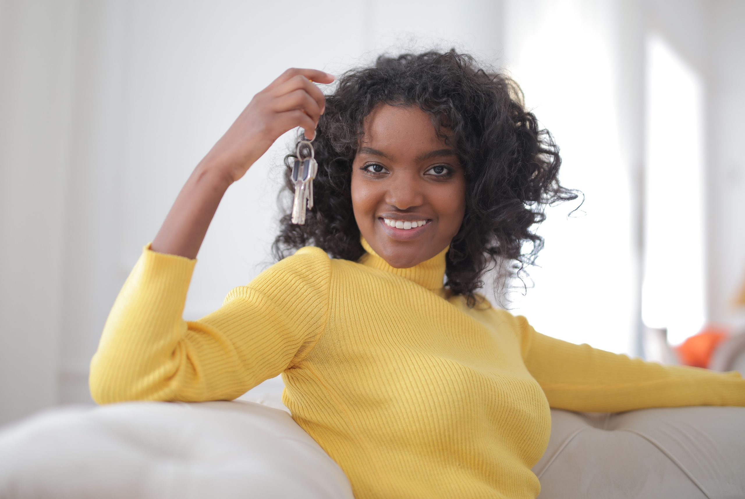 African American woman smiling and holding keys, sitting indoors, symbolizing new home ownership. Femme afro-américaine souriante tenant des clés, assise à l'intérieur, symbolisant l'accession à la propriété d'une nouvelle maison.