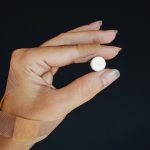 A hand wrapped in bandages holding a white pill against a dark background. Une main enveloppée de bandages tenant un comprimé blanc sur un fond sombre.