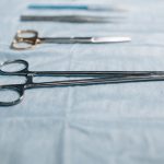 Close-up of surgical tools including a needle holder on a blue cloth in a medical setting. Gros plan sur des instruments chirurgicaux incluant un porte-aiguille sur un tissu bleu dans un contexte médical.