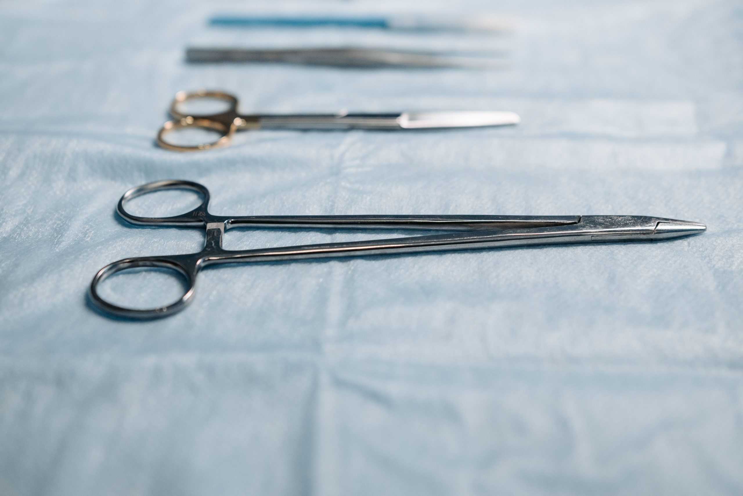 Close-up of surgical tools including a needle holder on a blue cloth in a medical setting. Gros plan sur des instruments chirurgicaux incluant un porte-aiguille sur un tissu bleu dans un contexte médical.