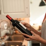 A senior woman examines a wine bottle in her cozy kitchen, wearing a striped apron. Une femme âgée examine une bouteille de vin dans sa cuisine confortable, portant un tablier rayé.