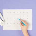 A hand marking a date on a calendar next to a glucometer on a purple background. Une main marquant une date sur un calendrier à côté d'un glucomètre sur un fond violet.