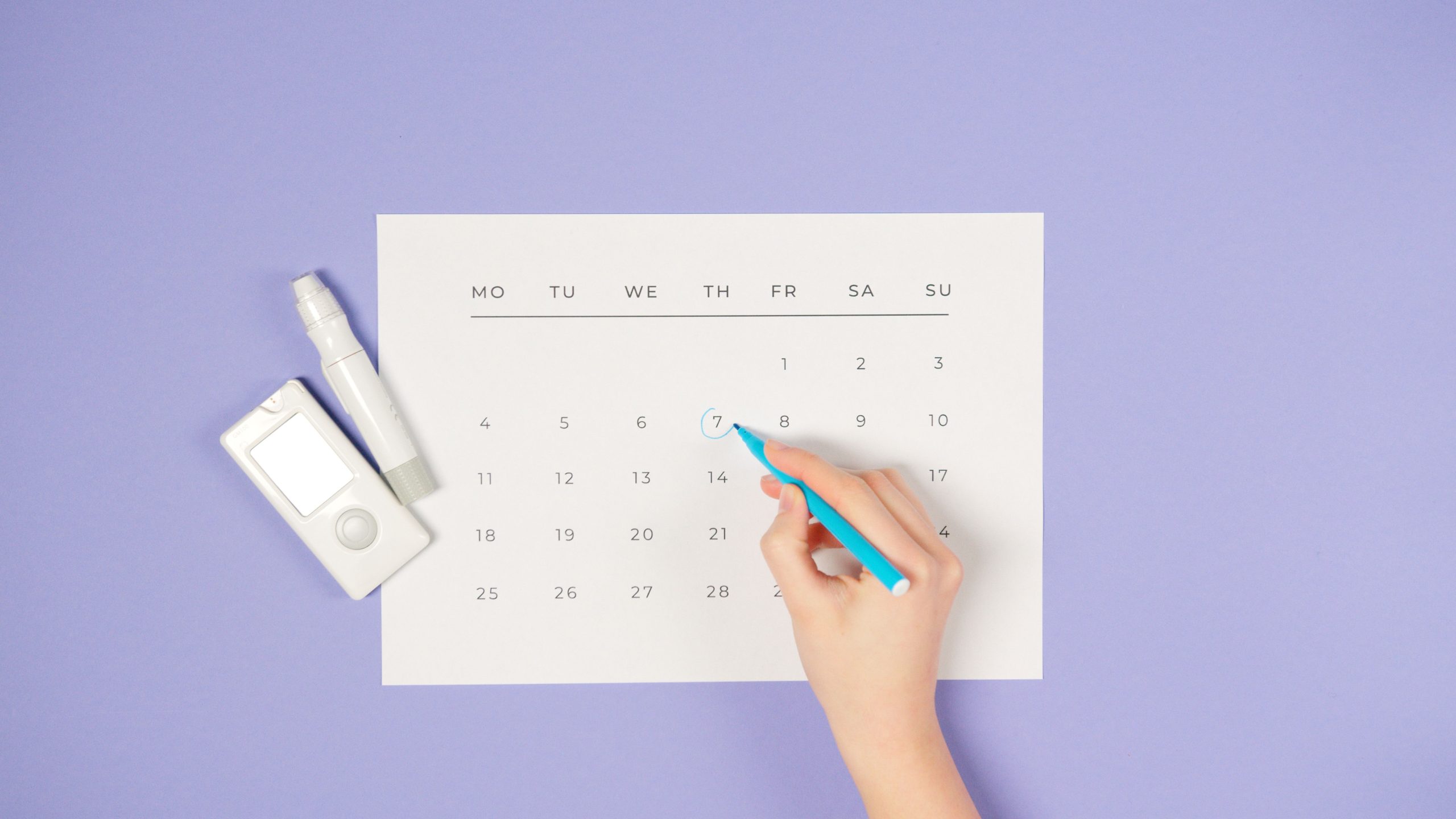 A hand marking a date on a calendar next to a glucometer on a purple background. Une main marquant une date sur un calendrier à côté d'un glucomètre sur un fond violet.