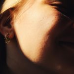 Close-up image capturing a woman smiling with sunlight highlighting her cheek and gold earring. Gros plan capturant une femme souriante avec la lumière du soleil mettant en évidence sa joue et sa boucle d'oreille en or.
