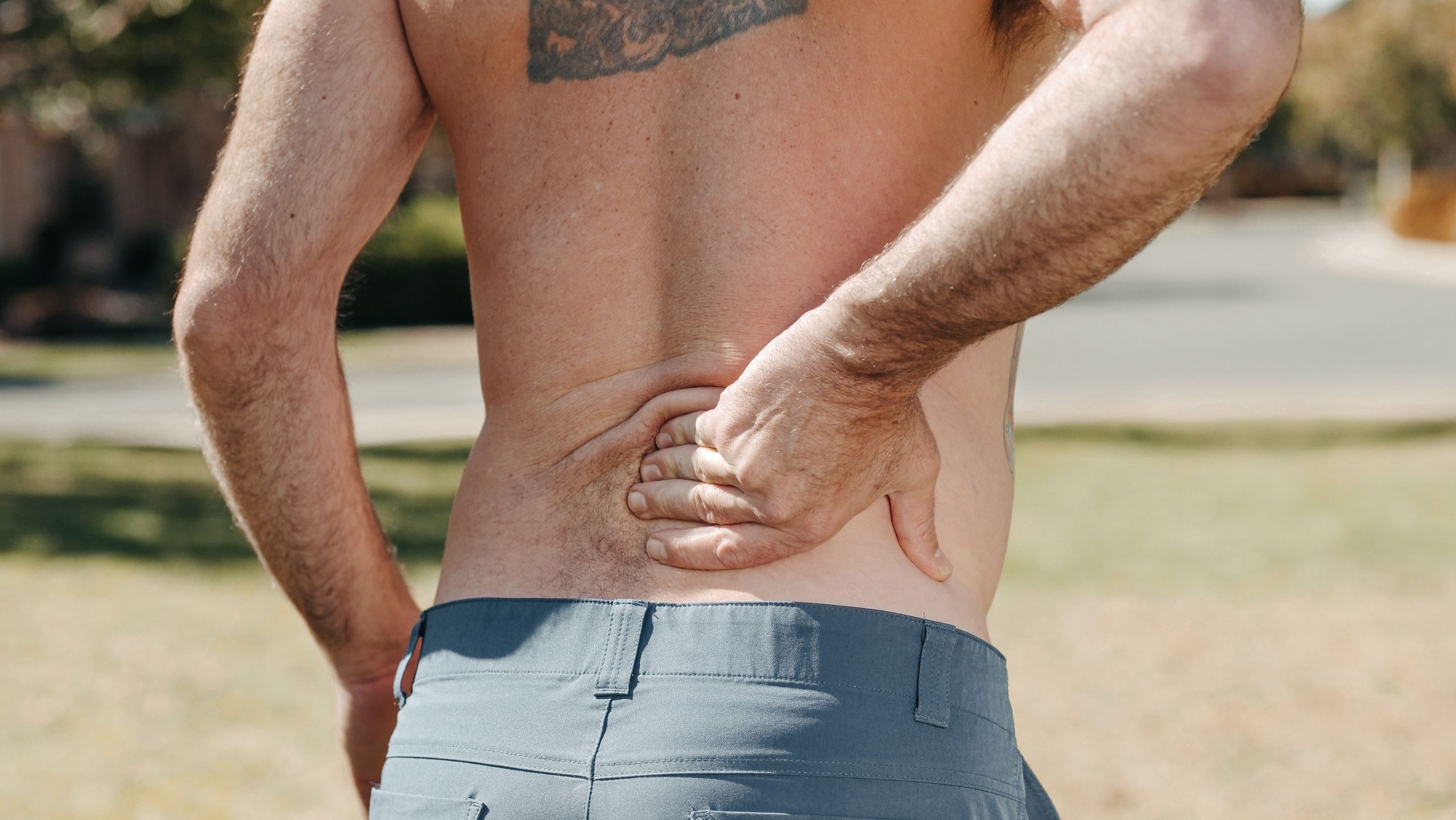 Shirtless man holding his lower back outdoors indicating pain or discomfort. Homme torse nu se tenant le bas du dos à l'extérieur indiquant une douleur ou un inconfort.