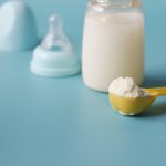 Close-up of baby milk formula powder in a yellow scoop with a bottle on a blue background. Gros plan de poudre de lait maternisé pour bébé dans une cuillère jaune avec une bouteille sur fond bleu.