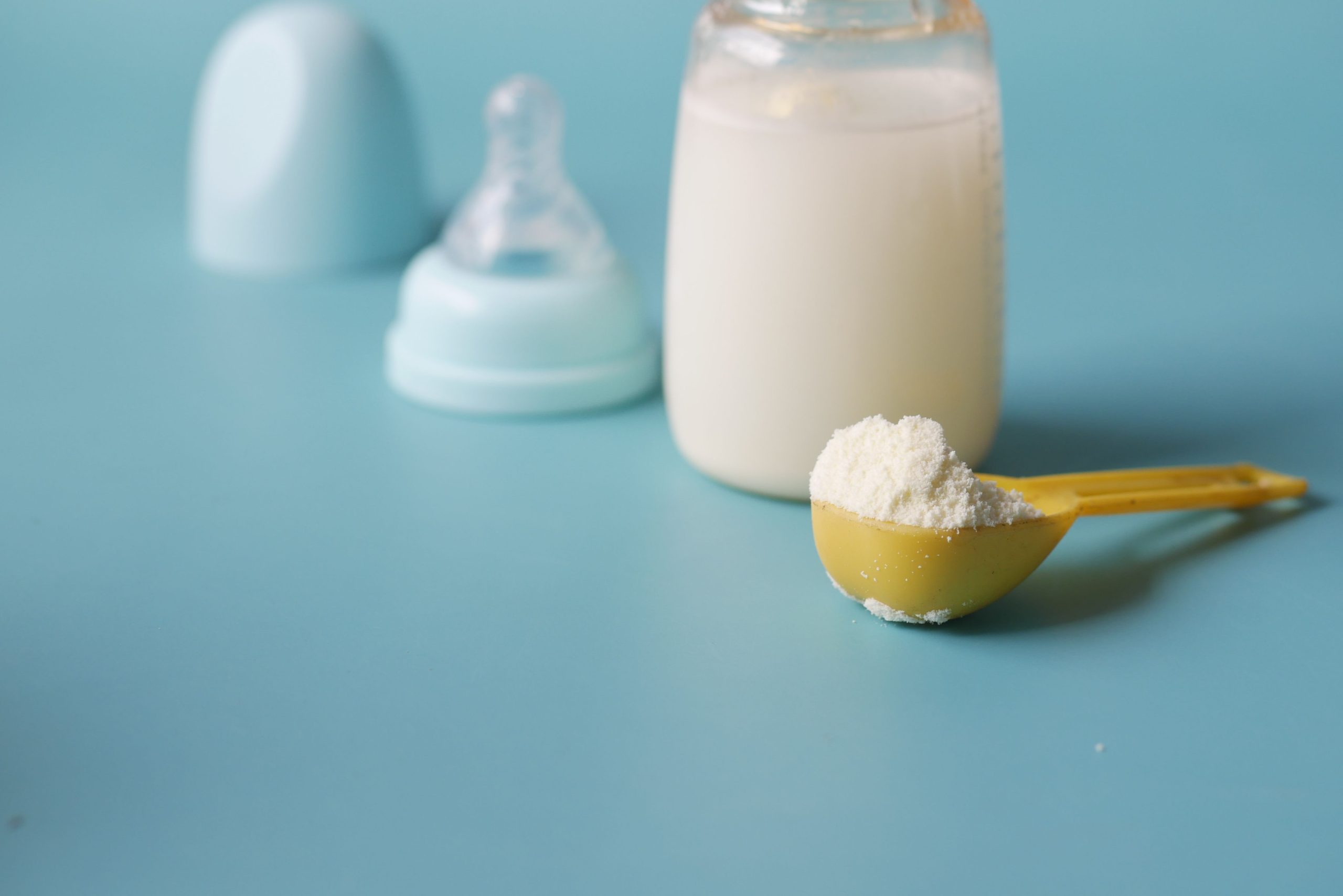 Close-up of baby milk formula powder in a yellow scoop with a bottle on a blue background. Gros plan de poudre de lait maternisé pour bébé dans une cuillère jaune avec une bouteille sur fond bleu.