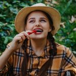 Fille souriante avec un chapeau de paille mangeant un poivron rouge à l'extérieur dans un cadre verdoyant.