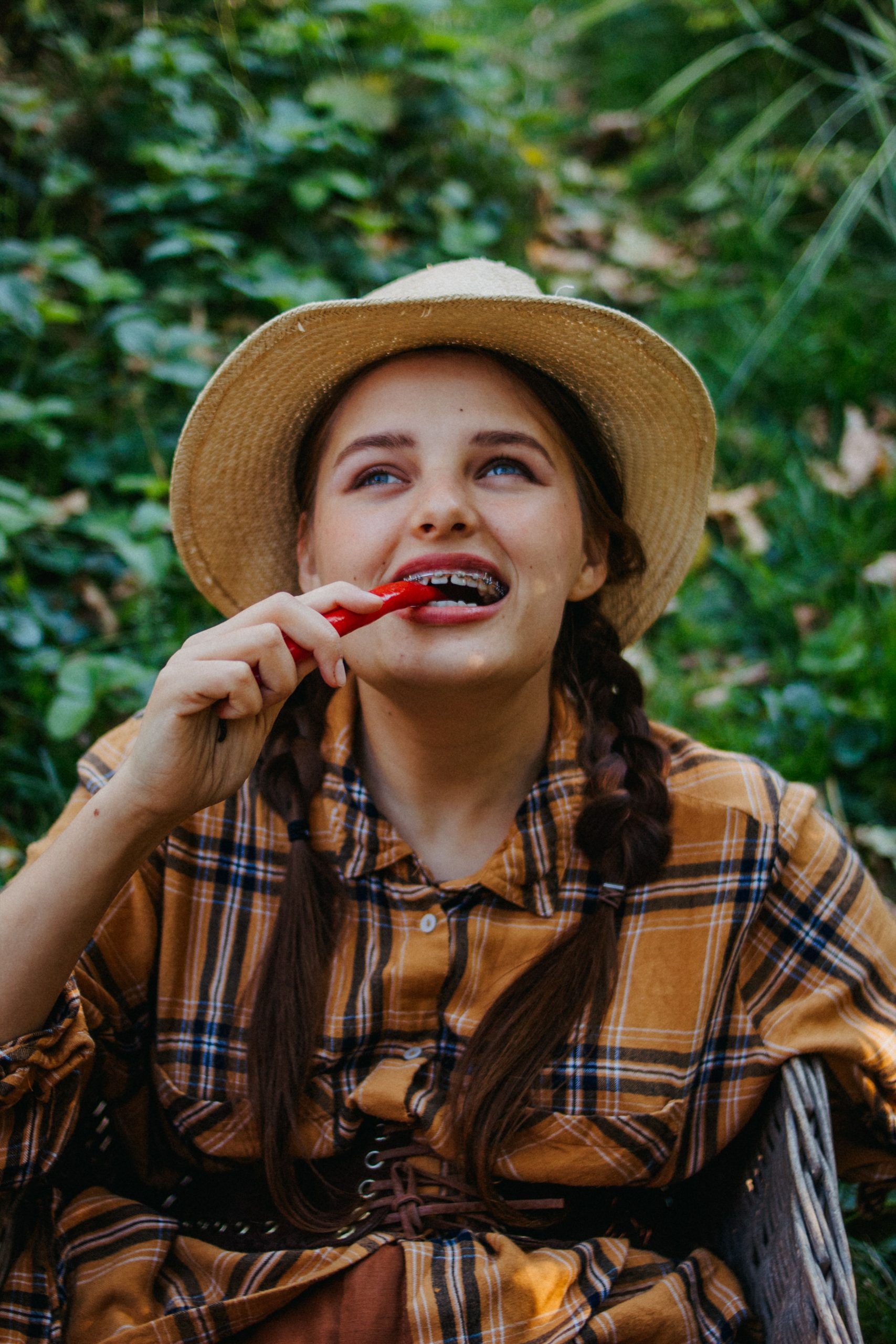 Fille souriante avec un chapeau de paille mangeant un poivron rouge à l'extérieur dans un cadre verdoyant.